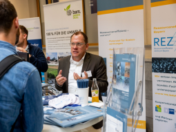 Am REZ-Stand im Ausstellerbereich unterhält sich die Person am Stand mit einem Teilnehmenden.